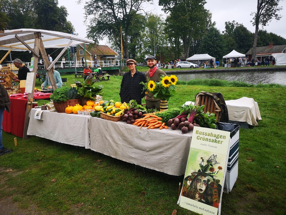 A market vegetable stand, two people behind a table filled with colorful vegetables and a sunflower bouquet